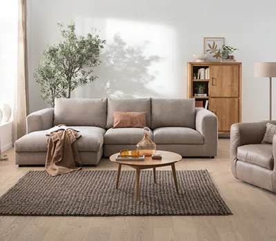 Helles Wohnzimmer mit großer beiger Wohnlandschaft, rundem Holztisch und Massivholzschrank, kombiniert mit neutraler Dekoration und viel natürlichem Licht für ein wohnliches Ambiente.